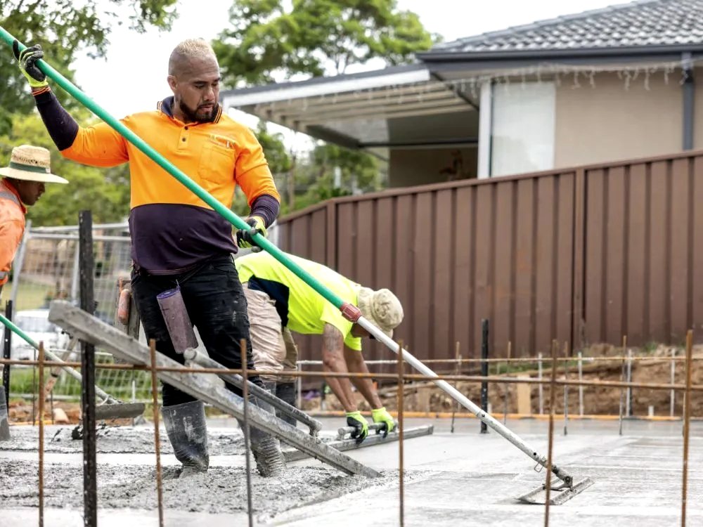 Two finishers screeding a freshly placed concrete slab beside a house